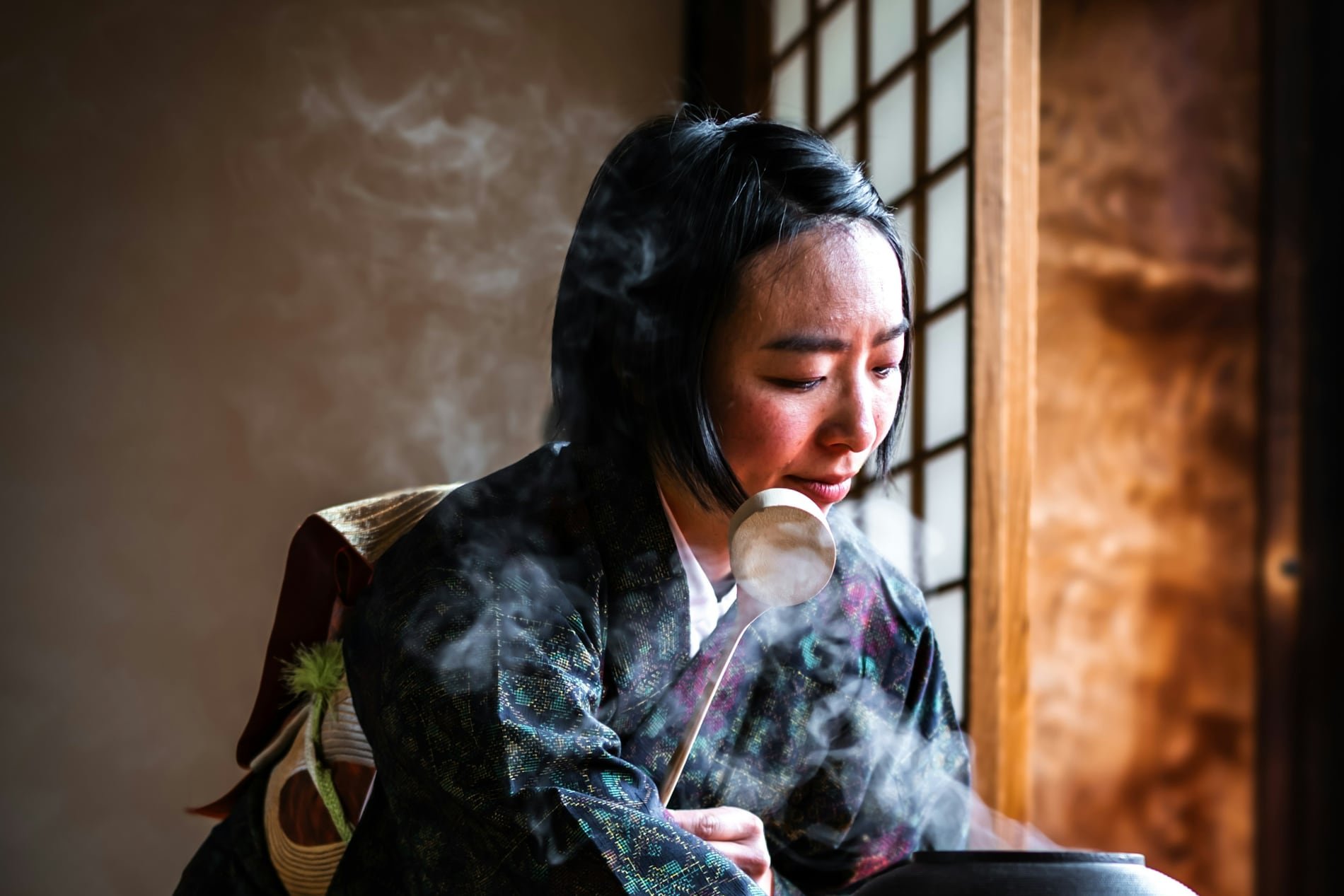 A woman in kimono preparing tea beside a steaming iron kettle