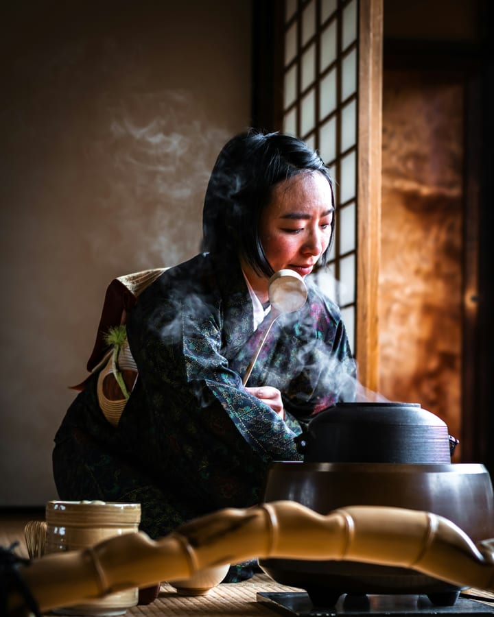 A woman in kimono preparing tea beside a steaming iron kettle